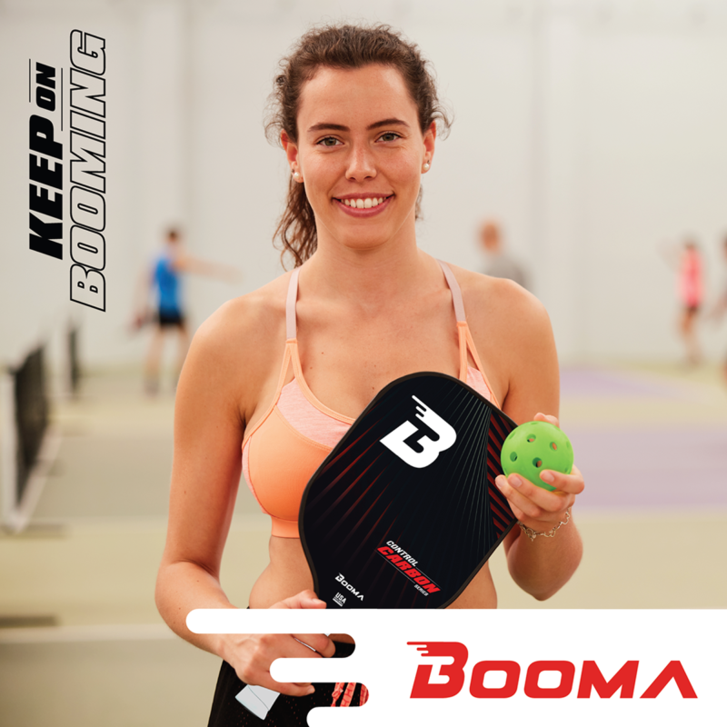 Jeune femme souriante avec sa raquette de pickleball BOOMA et sa balle sur la poitrine dans un gymnase.