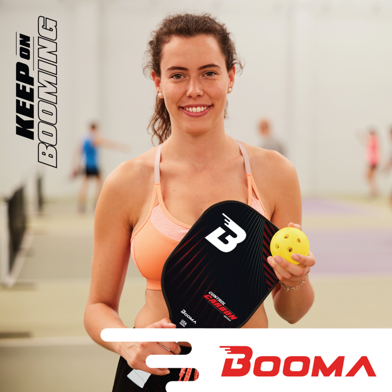 Jeune femme souriante avec sa raquette de pickleball BOOMA et sa balle sur la poitrine dans un gymnase.