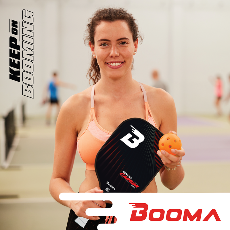 Young woman smiling with her BOOMA pickleball paddle and ball on her chest in a gym.